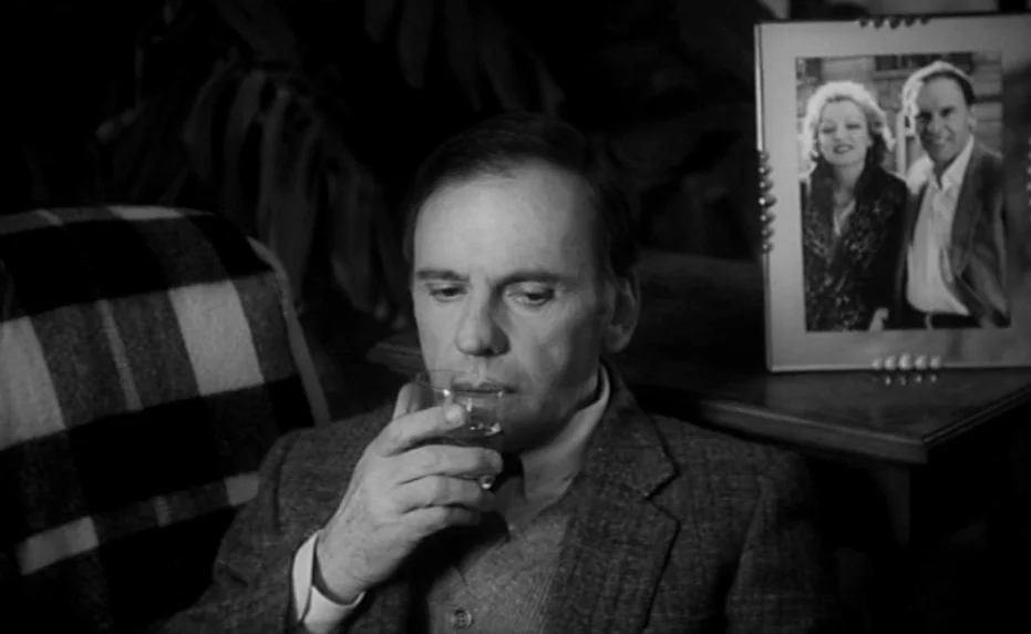 Vivement Dimanche (1983) movie still: medium — A man sits in a dimly lit room with a plaid blanket draped behind him. Next to him on a small table…
