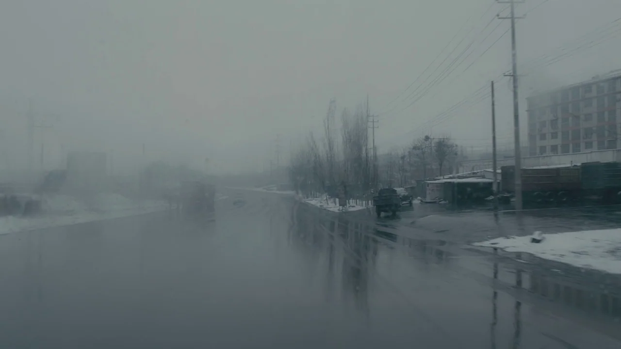 An Elephant Sitting Still (2018) movie still: wide — A wet asphalt road stretches through a foggy, industrial area during a light snowfall. Sparse…