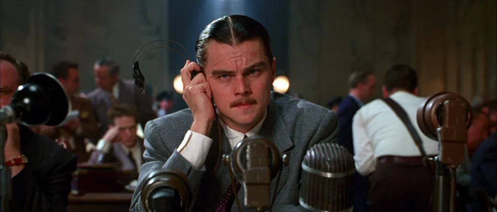 The Aviator (2004) movie still: medium — A crowded professional space with many men working behind microphones and desks. A man with a center-parted…