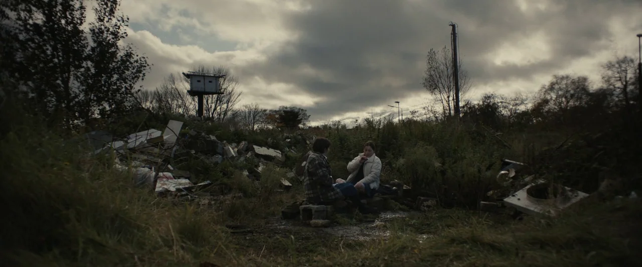 You Are Not My Mother (2021) movie still: wide — An overgrown vacant lot filled with piles of concrete rubble and debris under clouds. Two teenagers…