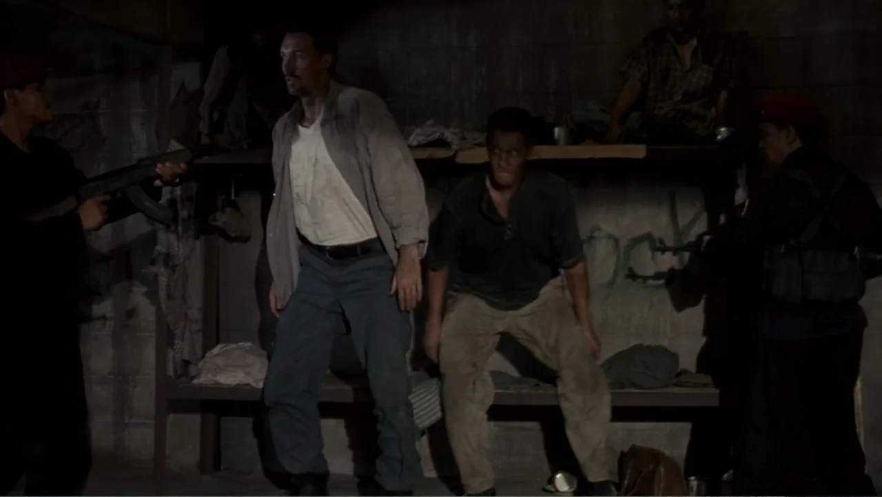 Behind Enemy Lines (2001) movie still: fullbody — A dimly lit cinderblock cell contains two men standing near a metal bed frame. Armed soldiers…