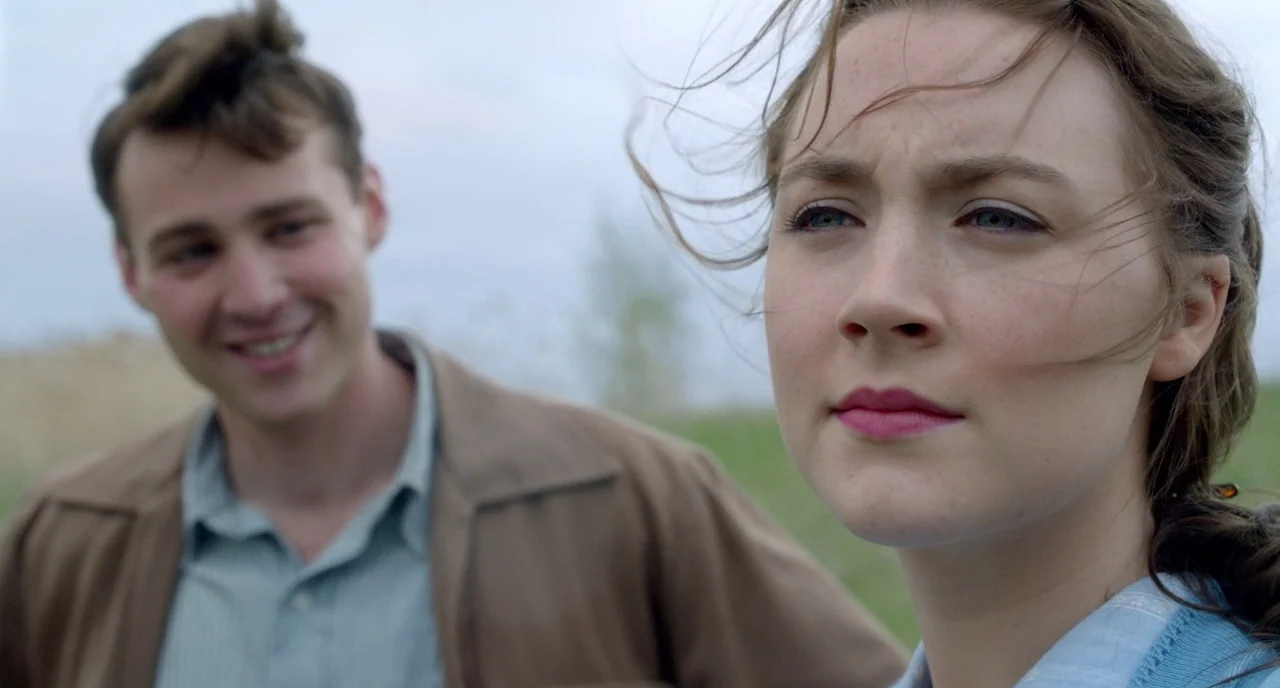 Brooklyn (2015) movie still: medium — A woman stands in a green grassy field under a pale overcast sky. A man looks at her from the background while…
