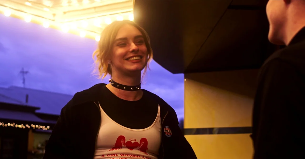 T Blockers (2023) movie still: medium — The exterior of a building at night features a glowing yellow marquee light strip. A young woman in a black…