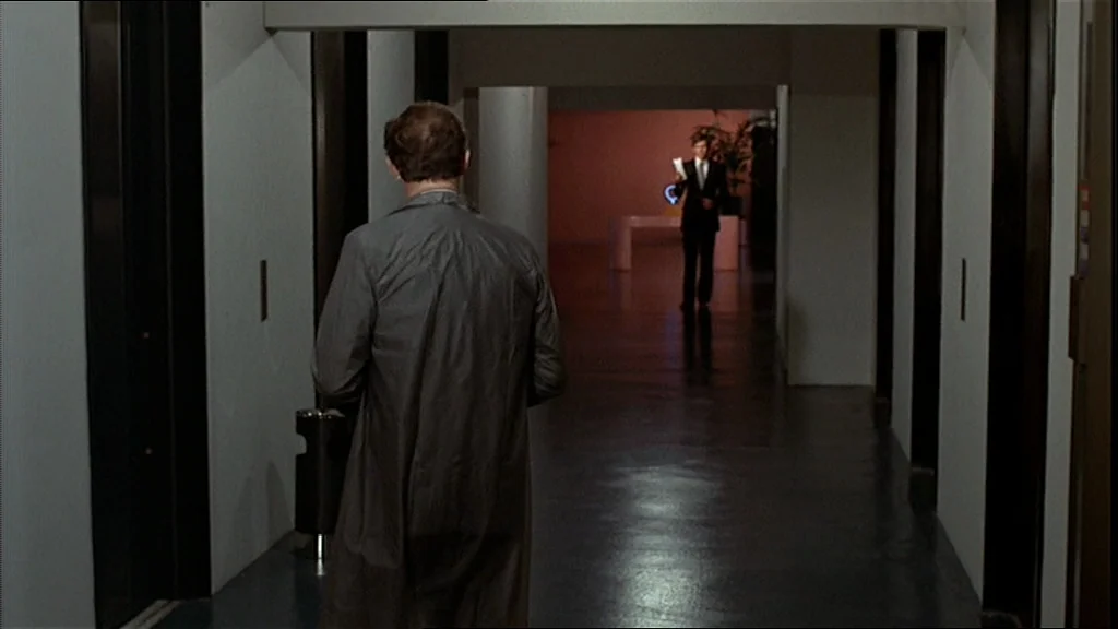 The Conversation (1974) movie still: medium — A long, sparsely decorated office corridor features dark trim and a smooth dark floor. Two men stand…