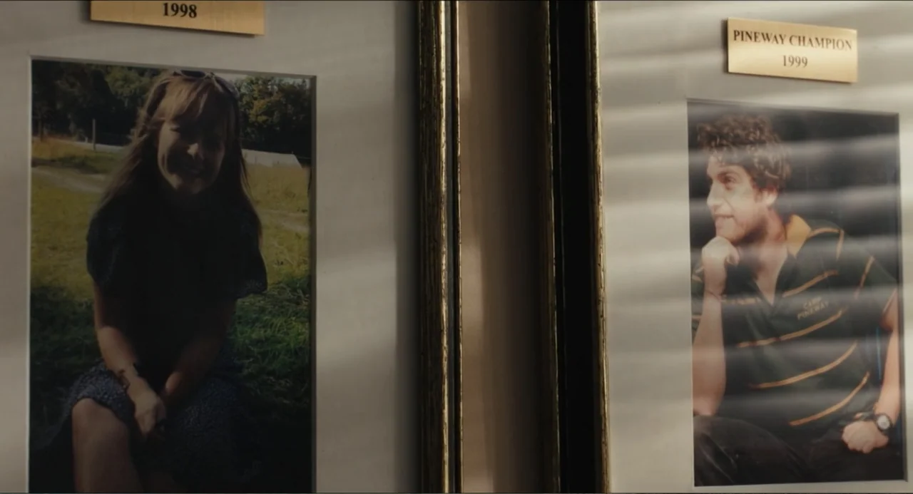 Hell of a Summer (2025) movie still: medium — Two framed photographs are mounted side-by-side on a light-colored wall with gold plaques above them.…