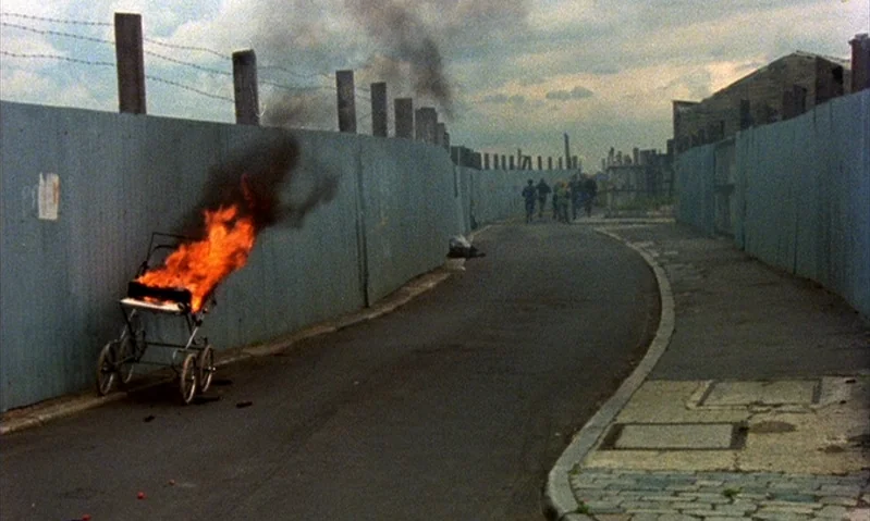 Jubilee (1978) movie still: wide — An urban alleyway is enclosed by tall corrugated metal fences topped with barbed wire. A metal baby carriage is…