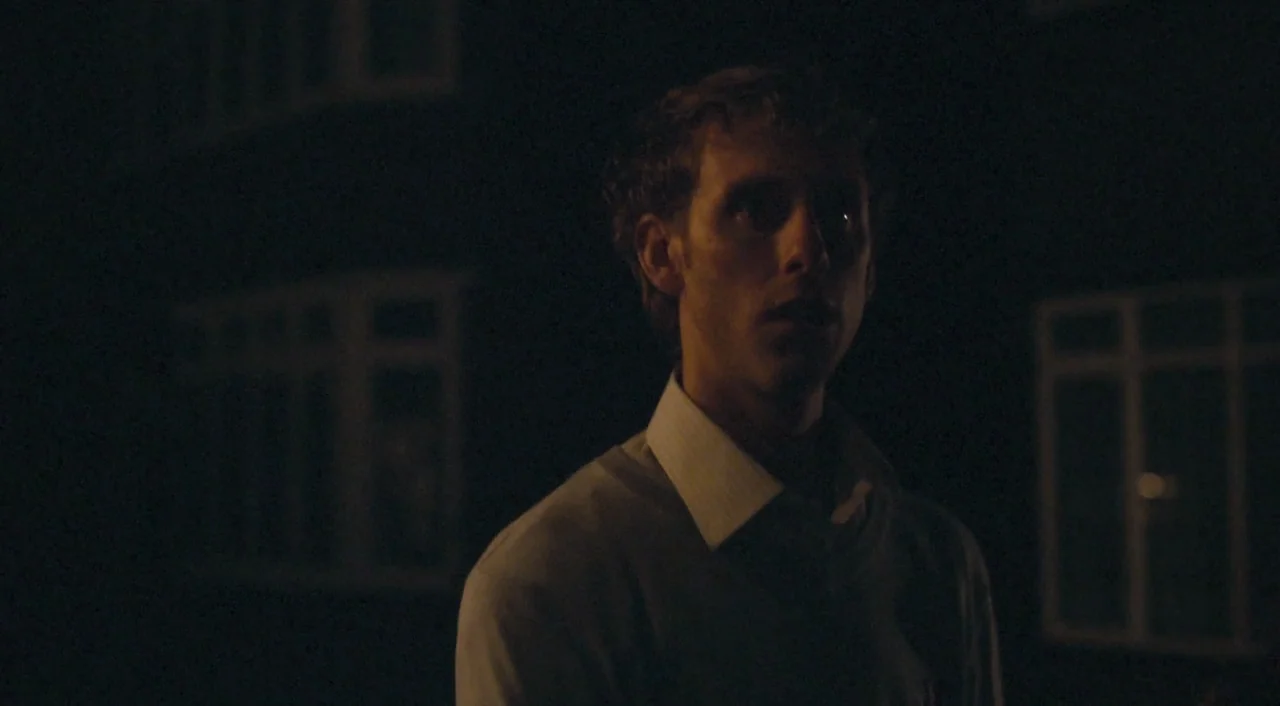 Broken (2012) movie still: medium — A man stands outdoors at night in front of a dark building with windows. He gazes off-camera while bathed in…