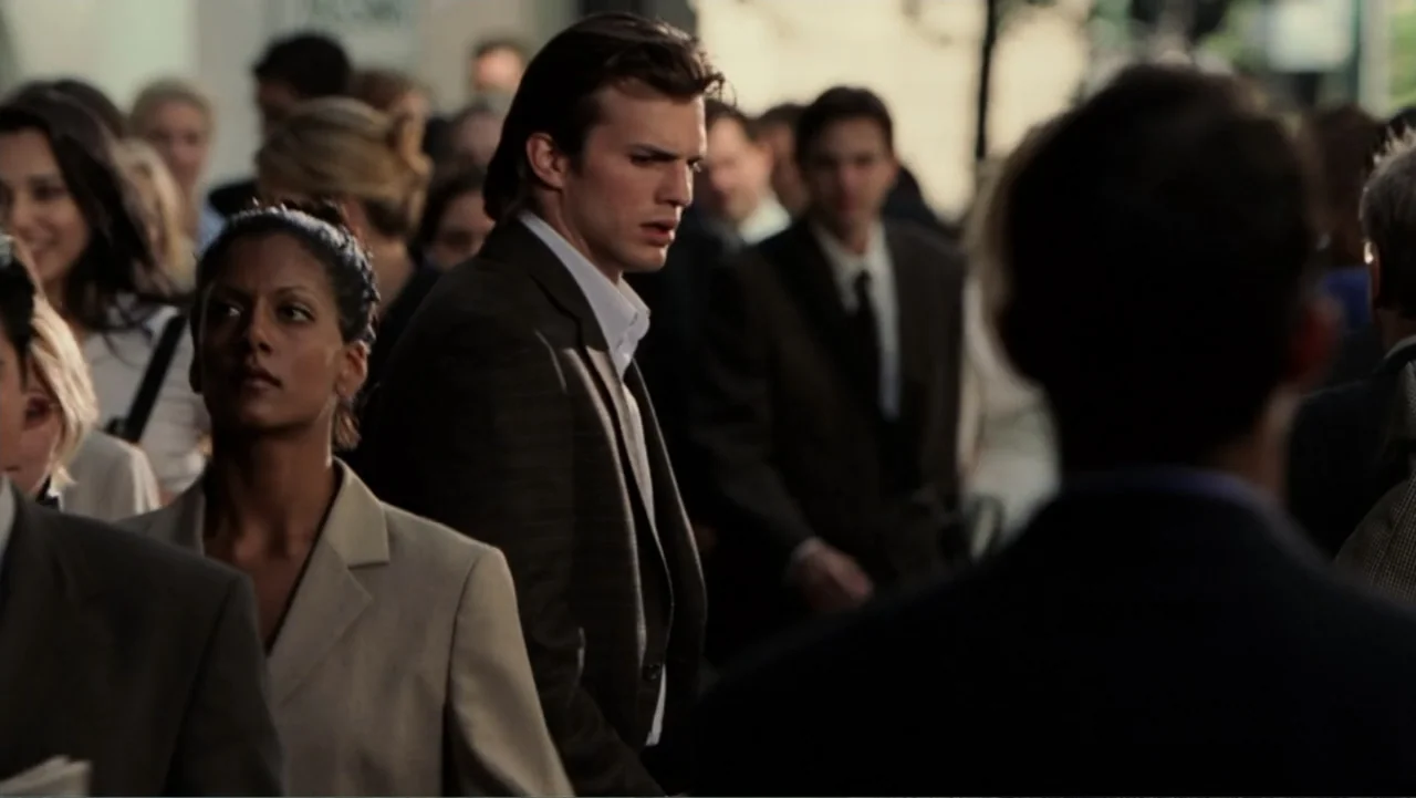 The Butterfly Effect (2004) movie still: medium — A dense urban crowd walks along a sidewalk during the day. A man in a dark blazer moves through…