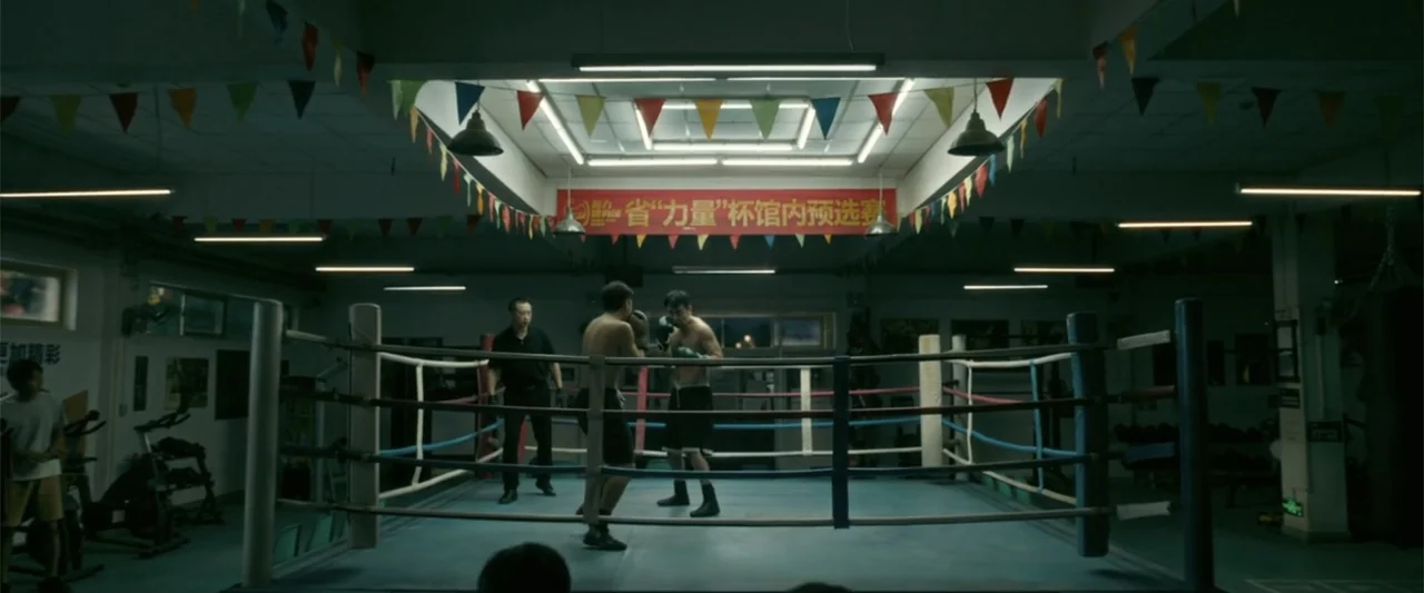 Yolo (2024) movie still: medium — A gymnasium interior features a raised boxing ring draped with festive triangular bunting flags. Two men spar…