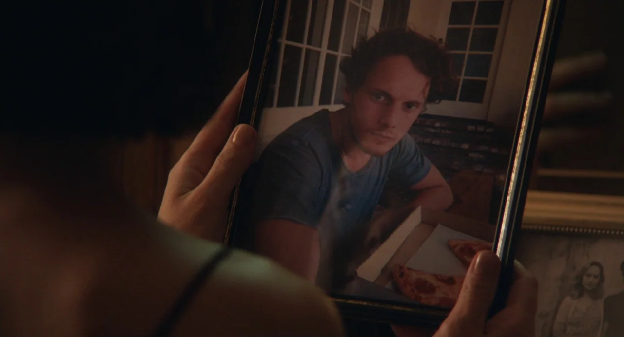 Cymbeline (2014) movie still: closeup — A person holds a black-framed photograph showing a young man sitting at a table. The man in the photo wears…