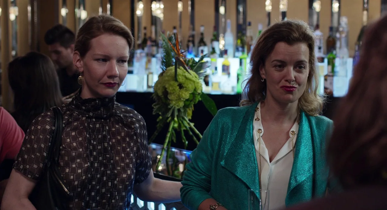 Toni Erdmann (2016) movie still: medium — A dimly lit bar with a backlit shelf of liquor bottles and a floral arrangement. Two women stand at the…