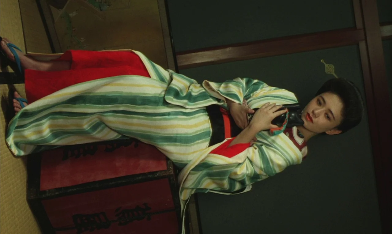 Yumeji (1991) movie still: fullbody — A traditional room features tatami mats and dark wall panels. A woman lies on a red lacquered chest while…
