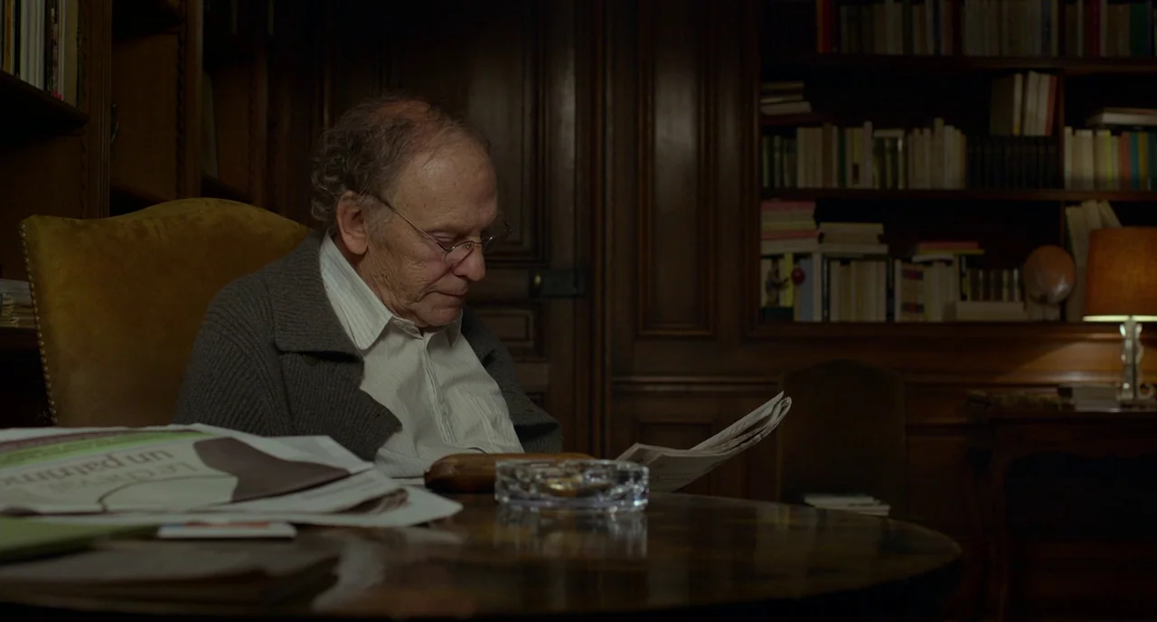 Amour (2012) movie still: medium — A dark, wood-paneled room is lined with shelves filled with numerous books. An elderly man with glasses sits in a…
