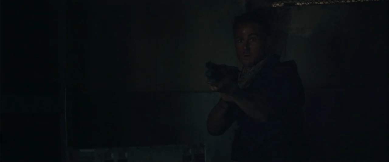 The Condemned 2 (2015) movie still: medium — A dark, dimly lit interior space with concrete walls and shadowed corners. A man in a tactical vest…