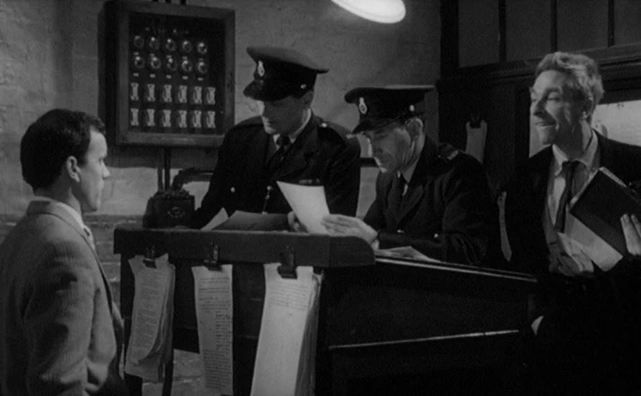 The Criminal (1960) movie still: medium — A utilitarian office space with rough stone walls and a tall, slanted desk. Two police officers review…