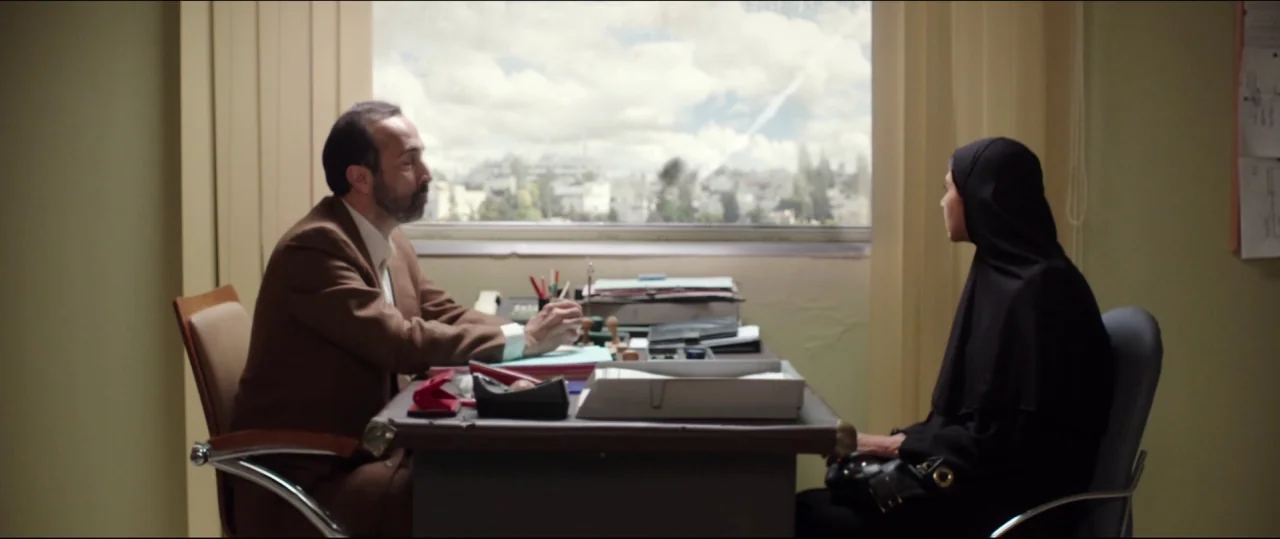 Under The Shadow (2016) movie still: medium — An office with olive-green walls faces a window overlooking a bright, cloudy cityscape. A man in a…