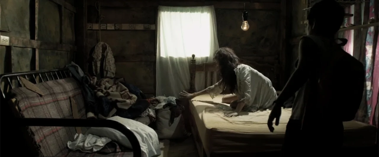 At Dawn (2013) movie still: medium — A dimly lit room features weathered wooden walls and a window with white curtains. A woman climbs onto a bare…