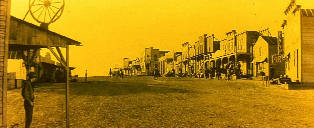 Lemonade Joe (1964) movie still: wide — A dusty main street of a wooden frontier town features a large wagon wheel sign. Multiple figures on…