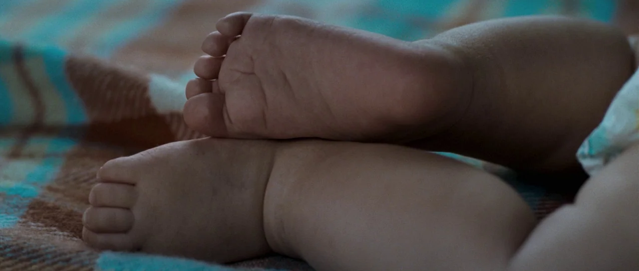 Womb (2010) movie still: closeup — A pair of infant feet rests on a woven wool blanket with a plaid pattern. The tiny legs are crossed and…
