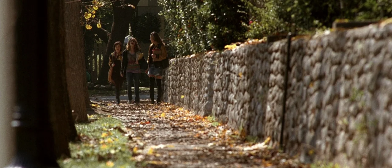 Halloween (2007) movie still: wide — A stone-walled sidewalk is scattered with dry orange leaves under a canopy of trees. Three young women walk…