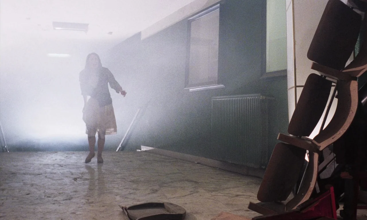 Demons (1985) movie still: fullbody — A long hallway has grey walls, a radiator, and thick white smoke filling space. A woman walks toward the…