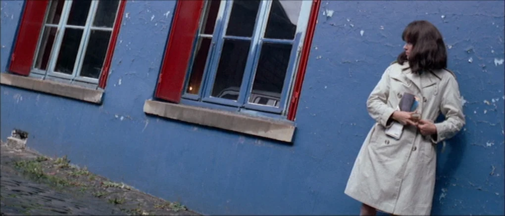 Made in U.S.A. (1966) movie still: fullbody — A woman stands against a bright blue wall with red window shutters outside. She rests against the…