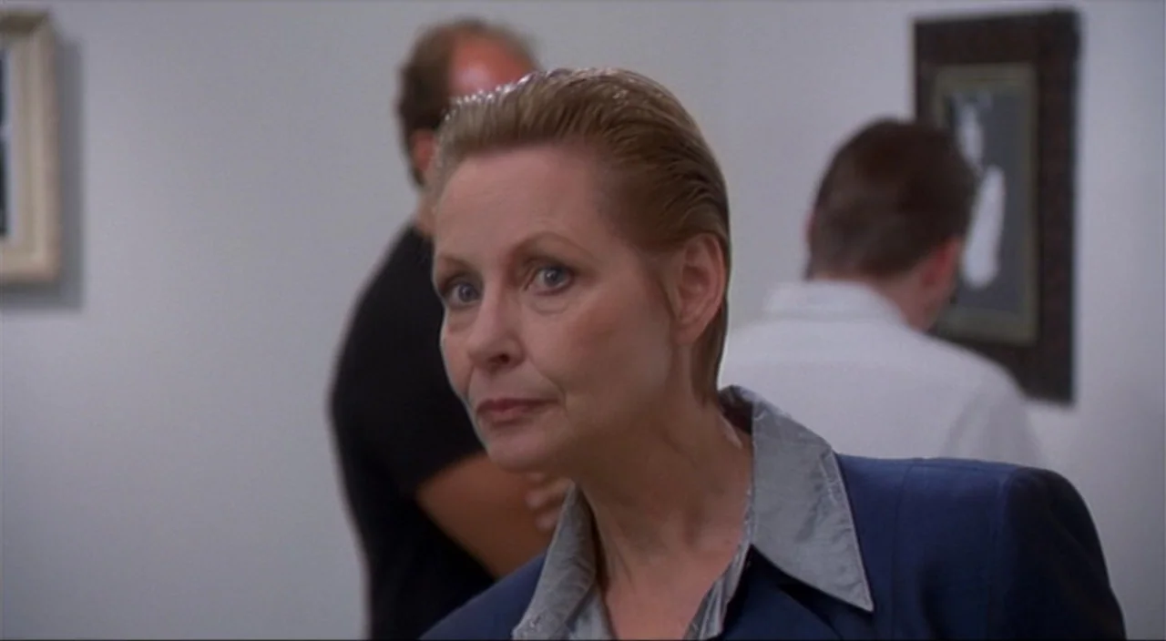 Pecker movie still: closeup — A brightly lit white room contains framed artworks hung on plain walls. A woman in a navy blazer stands foreground…