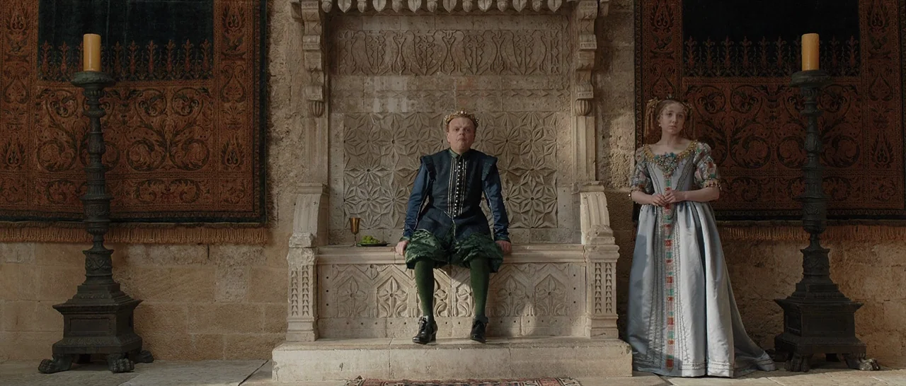 Tale of Tales (2015) movie still: wide — A stone chamber features an intricate carved throne flanked by two tall, dark candle stands. A man in…