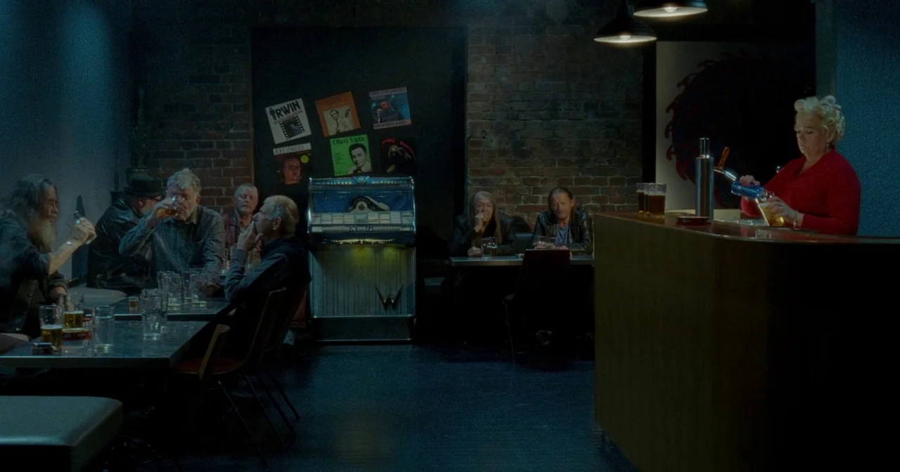 Fallen Leaves (2023) movie still: wide — A dimly lit bar features a brick wall decorated with album covers and music posters. Patrons sit at metal…