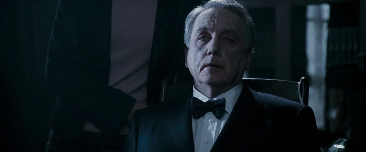 Dead Silence (2007) movie still: medium — A dimly lit indoor space with a person in formal wear sitting in a chair. An indistinct figure stands…