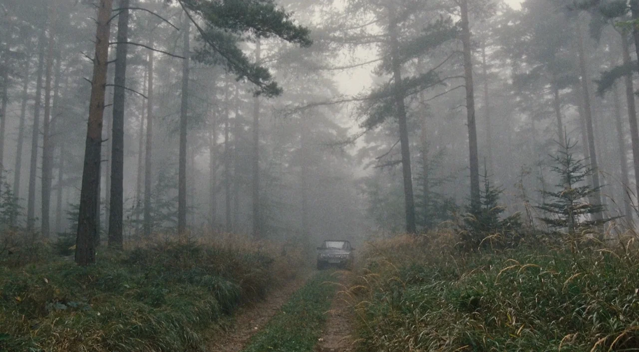Paradise: Hope (2013) movie still: wide — A dense coniferous forest is filled with thick ground fog and tall pine trees. A dark car drives slowly…