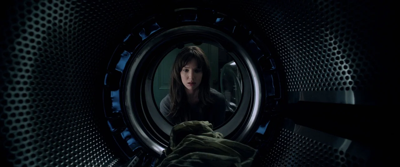 Malignant (2021) movie still: medium — A woman stares into the open metal drum of a dark clothes dryer. The machine's perforated steel interior…