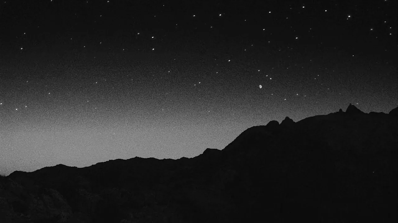 Divinity (2023) movie still: establishing — A dark rocky mountain range sits under a vast, clear night sky. Countless stars are scattered across the…