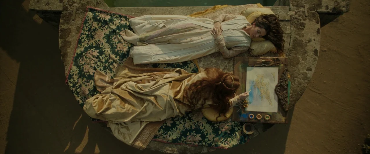 Rosaline (2022) movie still: fullbody — Two women rest on a patterned rug atop a stone structure near water. One woman is painting a map with…