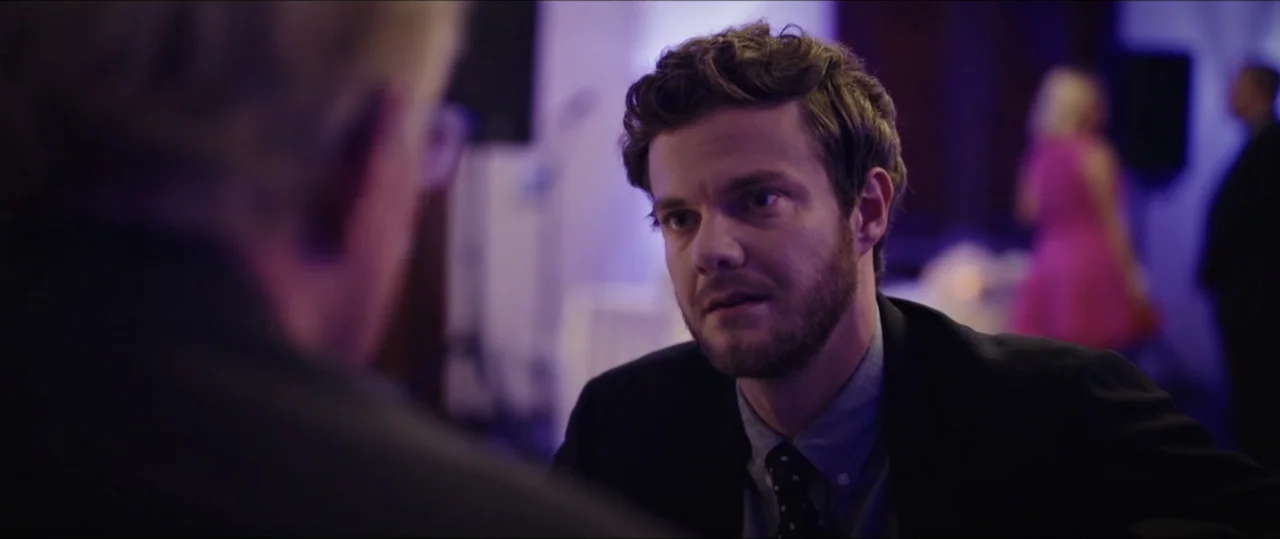 Plus One (2019) movie still: medium — A dimly lit indoor space with purple ambient lighting in the background. A man in a suit looks toward another…