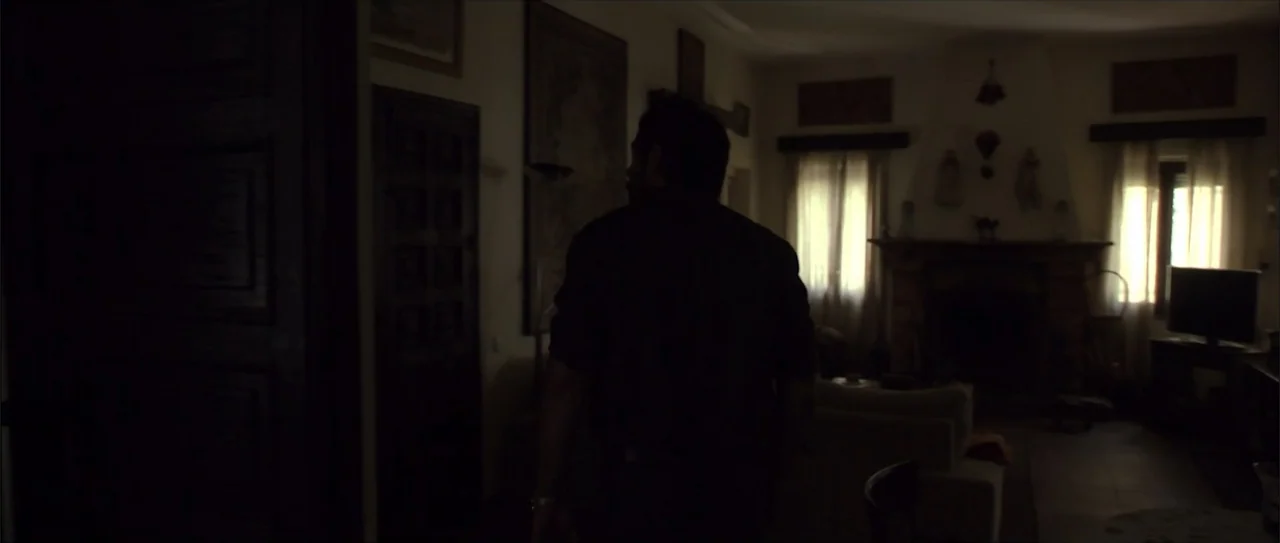 Otro verano (2013) movie still: medium — A dimly lit room with a fireplace and windows during the day. A man walks through a doorway toward the…