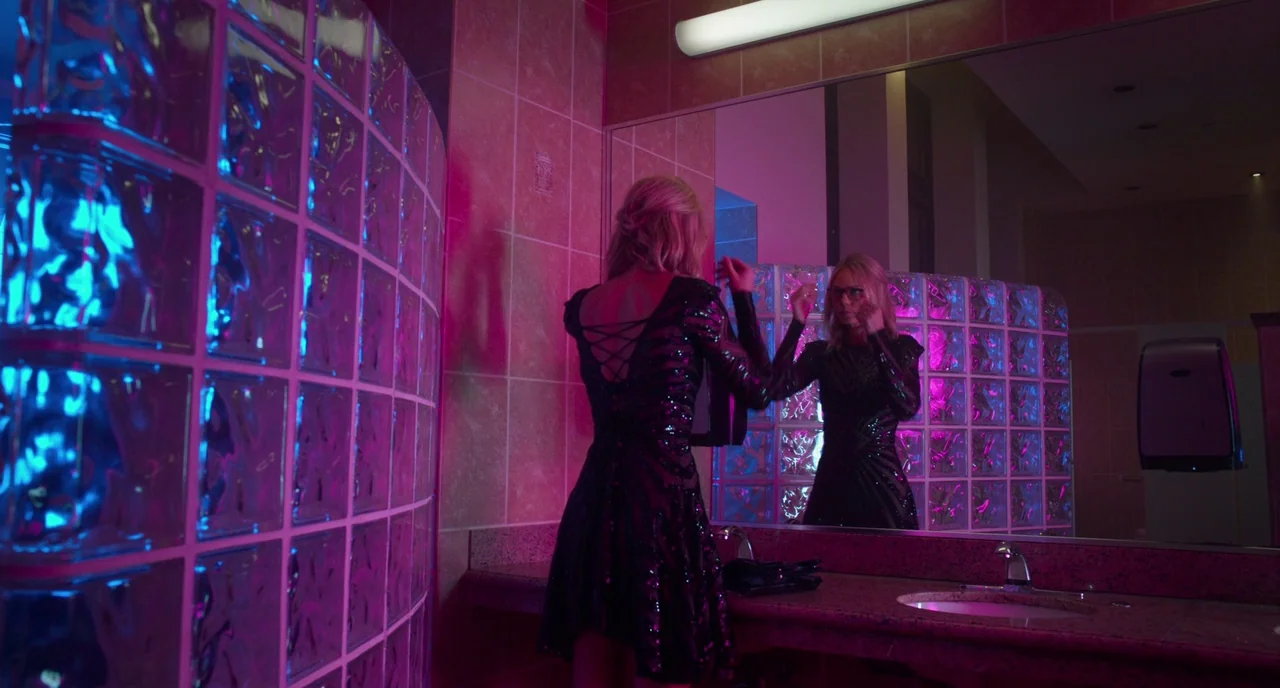 Rabid (2019) movie still: medium — A modern restroom with wall-to-wall glass block partitions illuminated by neon lighting. A woman stands at the…