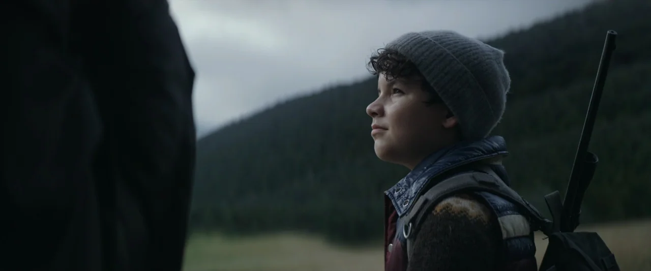 Wolf Man (2025) movie still: medium — A mountain side covered in dense forest under a heavy overcast gray sky. A young boy in a beanie and backpack…