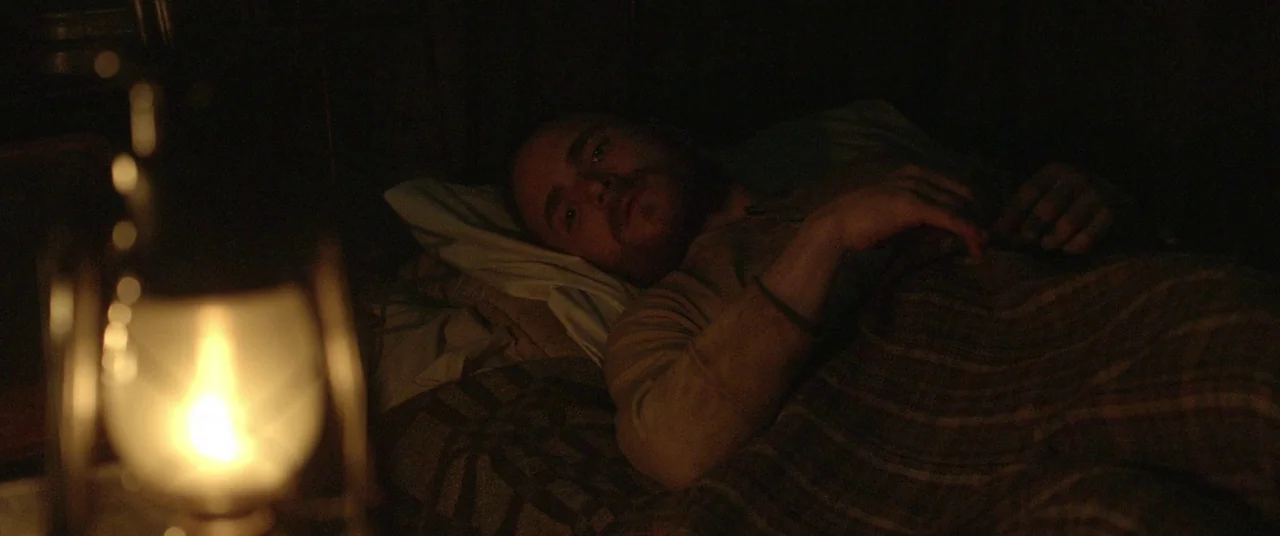 The Survivalist (2015) movie still: medium — A man lies in a dimly lit space with a textured blanket covering him. An oil lantern burns brightly in…