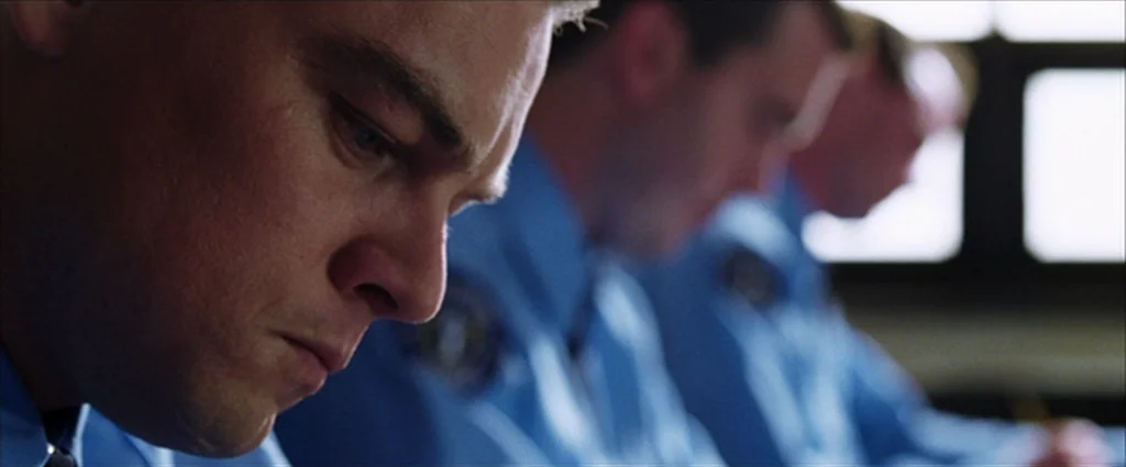 The Departed (2006) movie still: closeup — A group of police officers sit in a row within a bright classroom. They focus downward on their work…