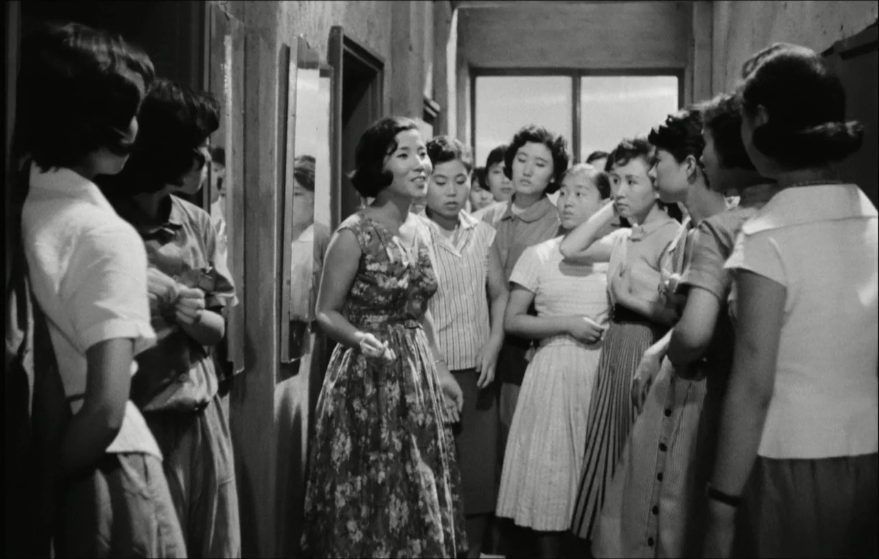 The Housemaid (1960) movie still: medium — A narrow school hallway with plain walls houses a group of young women. One woman stands in the center…