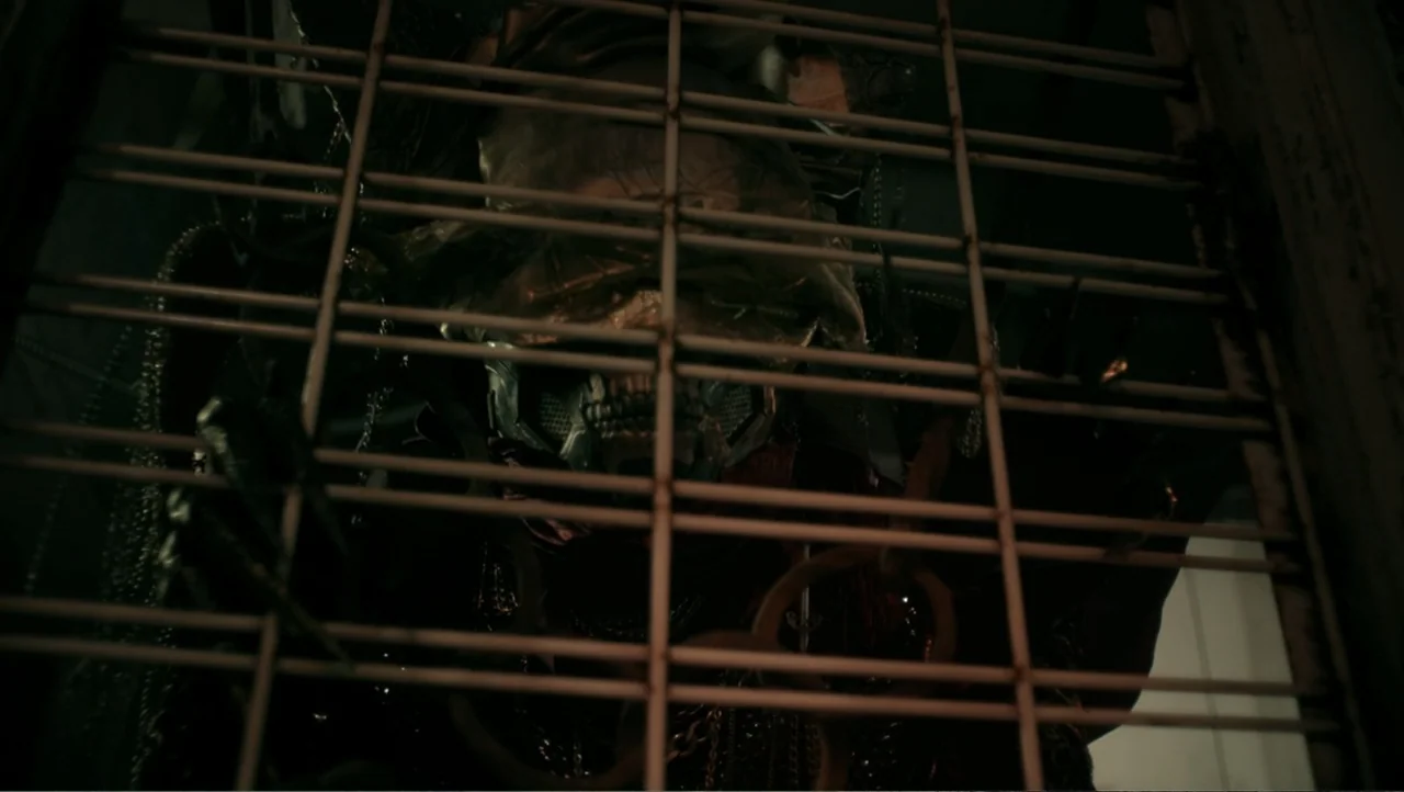 Shadow Master (2022) movie still: closeup — A dark, cramped space is partitioned by a heavy metal grid structure. A person wearing an elaborate…