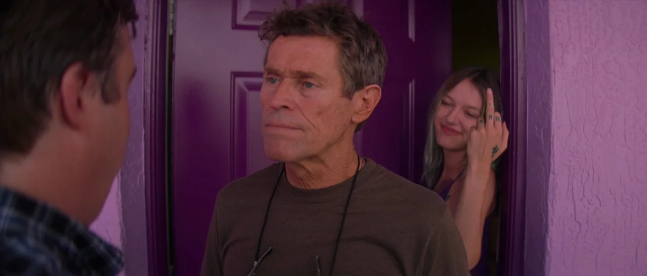 The Florida Project (2017) movie still: medium — A man stands before a vibrant purple door at a bright pink stucco motel. Behind him, a woman smiles…