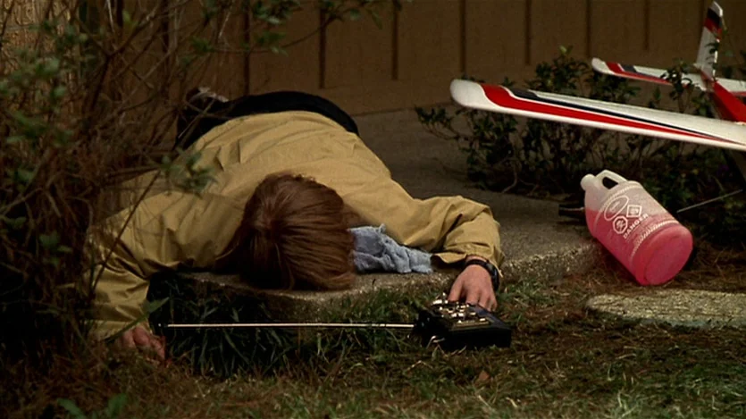 Love Liza (2002) movie still: medium — A person lies prone on a concrete edge near a suburban-style wall. They are positioned beside a small model…