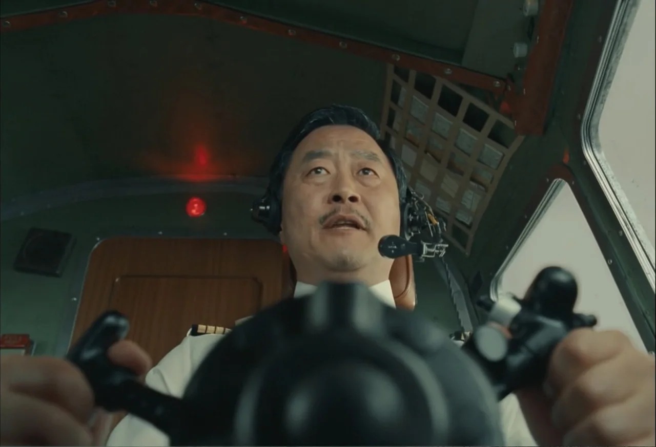 The Phoenician Scheme (2025) movie still: medium — The interior of an aircraft cockpit features metallic green wall panels and a control yoke. An…