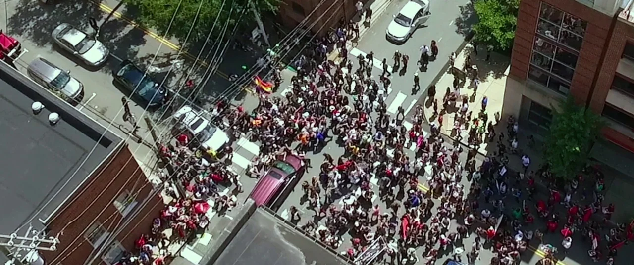 BlacKkKlansman (2018) movie still: wide — An aerial view of a dense crowd gathered at a street intersection during daytime. Many individuals are…