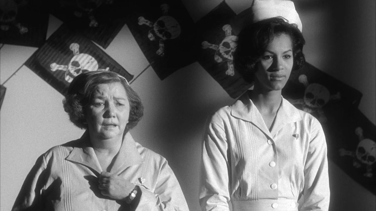 The Naked Kiss (1964) movie still: medium — A sterile interior wall features multiple framed skull and crossbones drawings hanging behind. Two…