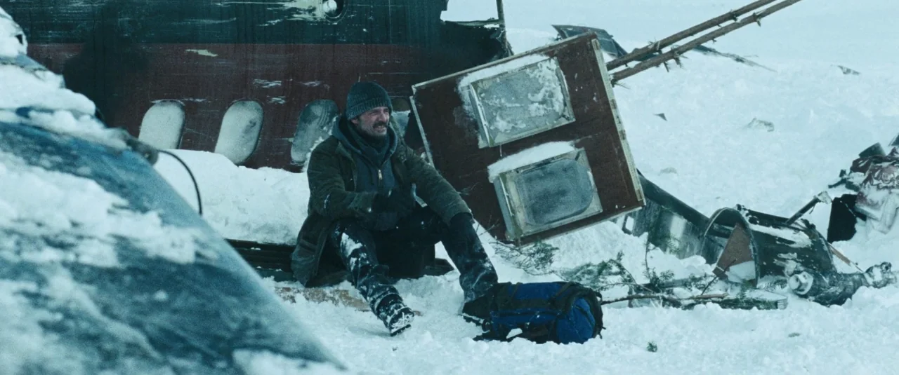 The Grey (2012) movie still: fullbody — A snowy, desolate location features the debris of a wrecked aircraft fuselage. A man in winter gear sits on…