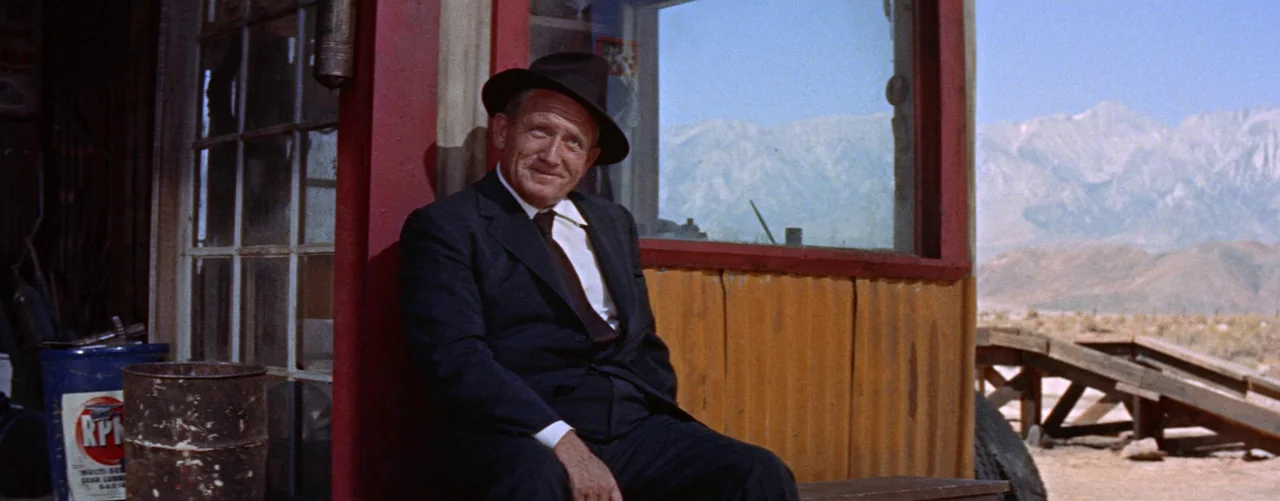 Bad Day At Black Rock (1955) movie still: medium — An aging man in a suit sits against a red-painted gas station exterior structure. In the…