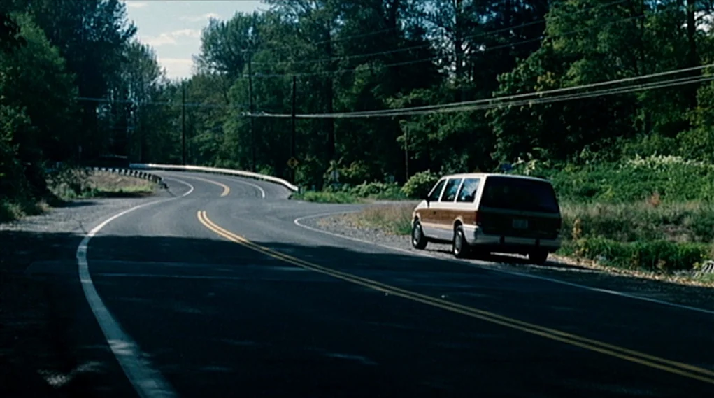 Visioneers (2008) movie still: wide — A curving paved country road winds through a dense line of green trees. A light-colored minivan with…